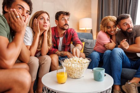 Foto de Grupo de amigos que se divierten pasando tiempo libre juntos en casa, comiendo palomitas de maíz y viendo películas de miedo en la televisión - Imagen libre de derechos