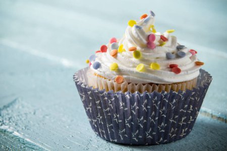Téléchargez les photos : Un muffin joliment décoré, avec crème et saupoudres colorées placées sur fond de planches de bois bleu clair - en image libre de droit