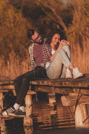 Foto de Hermosa pareja joven enamorada sentada en los muelles del lago, abrazándose y disfrutando de una hermosa puesta de sol sobre el lago - Imagen libre de derechos