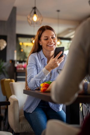 Foto de Mujer joven usando un teléfono inteligente y escribiendo mensajes de texto mientras está en una cita en un restaurante - Imagen libre de derechos