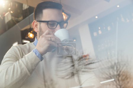 Photo for Portrait of a male freelancer sitting at a restaurant table, drinking coffee and working on a laptop computer - Royalty Free Image