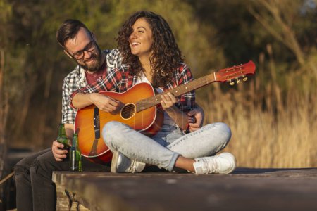 Photo for Couple sitting at the lake docks, playing the guitar, singing and drinking beer, enjoying a day in nature and relaxing - Royalty Free Image