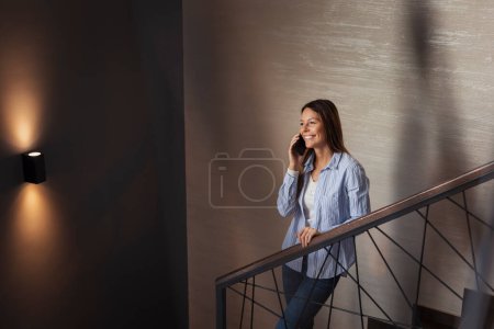 Photo for Beautiful young woman standing on a staircase, smiling while having a phone conversation - Royalty Free Image