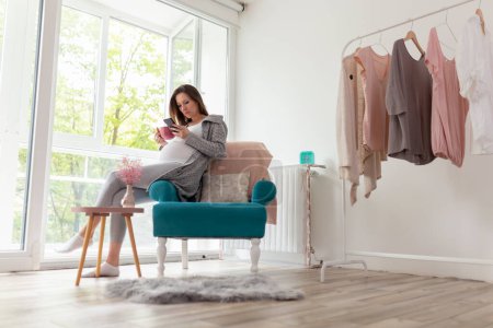 Téléchargez les photos : Femme enceinte assise dans un fauteuil, relaxante à la maison, buvant une tasse de thé chaud et utilisant un téléphone intelligent - en image libre de droit