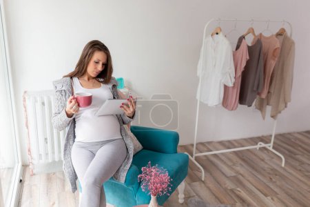 Foto de Hermosa mujer embarazada relajándose en casa, sentada en un sillón y leyendo blogs de maternidad en una tableta - Imagen libre de derechos