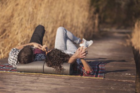 Foto de Pareja enamorada acostada en una manta de picnic en los muelles del lago, cogida de la mano, mirando al cielo y disfrutando de un hermoso día soleado de otoño al aire libre. - Imagen libre de derechos