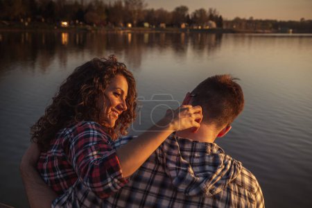 Foto de Hermosa pareja joven enamorada sentada en los muelles de un lago, abrazando y disfrutando de una hermosa puesta de sol sobre el lago y un día en la naturaleza - Imagen libre de derechos