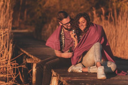 Téléchargez les photos : Couple assis sur les quais du lac, couvert de couverture, boire de la bière et du punch chaud et s'amuser - en image libre de droit