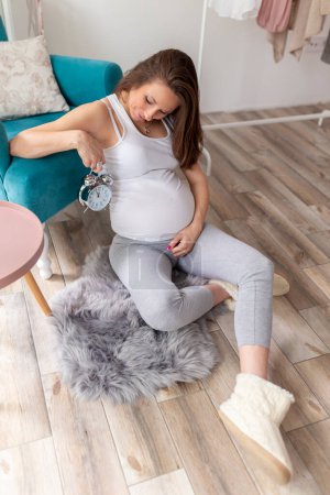 Téléchargez les photos : Jeune femme enceinte se détendant à la maison, attendant son bébé, tenant une horloge - compte à rebours pour une date d'échéance de livraison - en image libre de droit