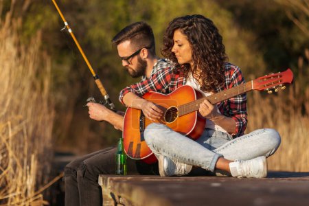 Téléchargez les photos : Couple assis sur les quais du lac, homme pêche pendant que la femme joue de la guitare, profiter d'une journée paisible dans la nature et se détendre - en image libre de droit