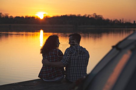 Téléchargez les photos : Jeune couple amoureux campant sur les quais du lac, assis sur le bord du quai à côté de la tente, embrassant et profitant d'un magnifique coucher de soleil sur le lac - en image libre de droit
