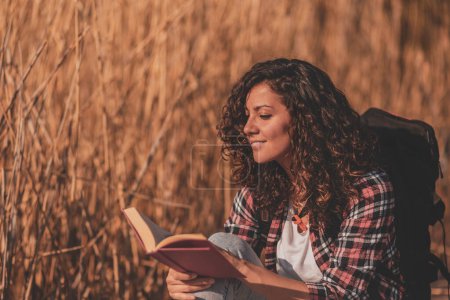 Téléchargez les photos : Belle jeune femme assise sur les quais du lac en bois, profitant d'une belle journée d'automne dans la nature et lisant un livre - en image libre de droit