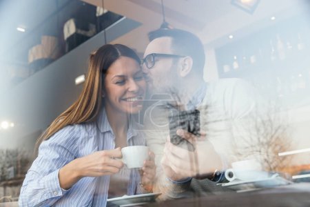 Portrait of a young couple in love sitting at a restaurant table, using a smart phone and kissing while drinking coffee