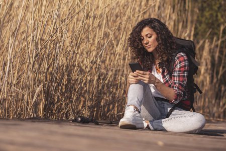 Téléchargez les photos : Femme assise sur les quais du lac en bois, profitant d'une journée d'automne dans la nature, se détendre après une randonnée et dactylographier un message texte sur un smartphone - en image libre de droit