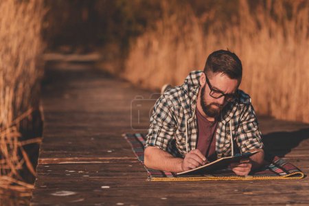 Téléchargez les photos : Homme profitant d'une journée ensoleillée d'automne dans la nature, dessinant des paysages dans un carnet de croquis - en image libre de droit