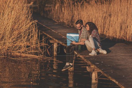 Téléchargez les photos : Couple assis sur les quais du lac, pêche et profiter d'une belle journée ensoleillée dans la nature - en image libre de droit