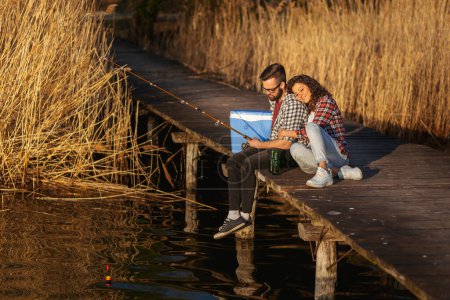 Téléchargez les photos : Couple assis sur les quais du lac, pêche et profiter d'une belle journée ensoleillée dans la nature - en image libre de droit