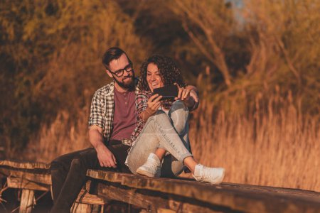 Téléchargez les photos : Couple assis sur les quais du lac, prendre un selfie avec un téléphone intelligent, boire de la bière et s'amuser - en image libre de droit