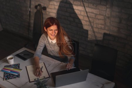 Téléchargez les photos : Vue en grand angle de la femme travaillant à distance de la maison, prenant des notes lors d'une conférence téléphonique à l'aide d'un ordinateur portable - en image libre de droit