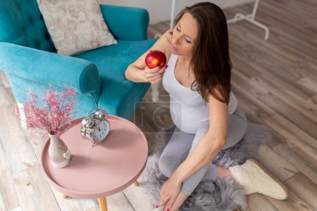 Foto de Vista de ángulo alto de una hermosa mujer embarazada sentada en el suelo junto a un sillón, relajándose en casa y comiendo una manzana - Imagen libre de derechos