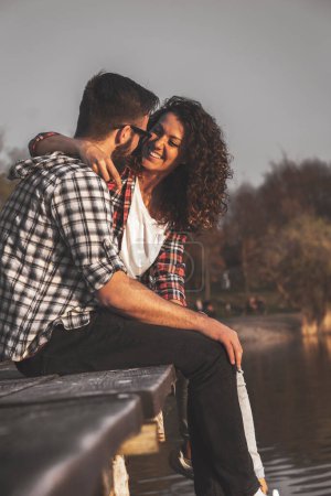 Foto de Hermosa pareja joven enamorada sentada en los muelles de un lago, abrazándose y disfrutando de una hermosa puesta de sol sobre el lago - Imagen libre de derechos
