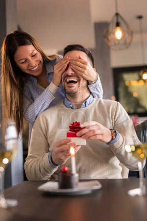 Téléchargez les photos : Couple à un rendez-vous au restaurant, célébrant l'anniversaire de l'homme ; femme tenant les yeux de l'homme et lui donnant un cadeau et un petit gâteau surprise - en image libre de droit