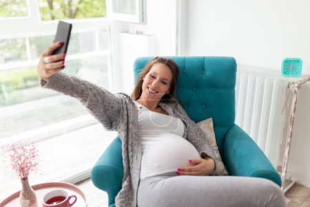 Téléchargez les photos : Femme enceinte relaxante à la maison, assise dans un fauteuil, buvant du thé et prenant des selfies avec un smartphone - en image libre de droit