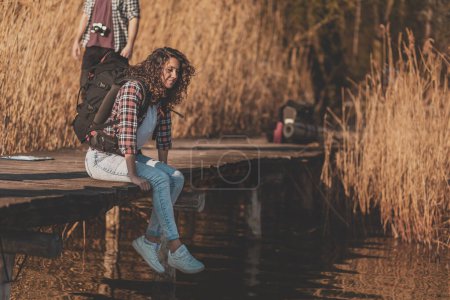 Téléchargez les photos : Deux routards faisant une pause, assis au bord du lac et relaxant, profitant d'une journée d'été ensoleillée dans la nature - en image libre de droit