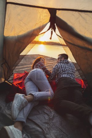 Téléchargez les photos : Jeune couple amoureux campant au bord du lac, allongé sur l'entrée de la tente, profitant d'un magnifique coucher de soleil - en image libre de droit