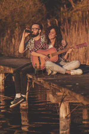 Téléchargez les photos : Couple assis sur les quais du lac, jouant de la guitare, chantant et buvant de la bière, profitant d'une journée dans la nature et se relaxant - en image libre de droit