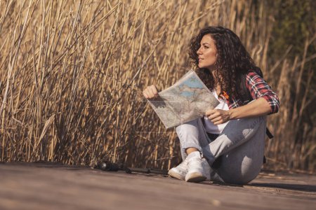 Téléchargez les photos : Femme assise sur les quais du lac en bois, prenant une pause de randonnée dans la nature, se détendre et lire une carte, planifier un itinéraire de randonnée - en image libre de droit