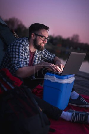 Foto de Hombre acampando junto al lago, sentado en la entrada de la tienda y trabajando en una computadora portátil; estilo de vida nómada digital - Imagen libre de derechos