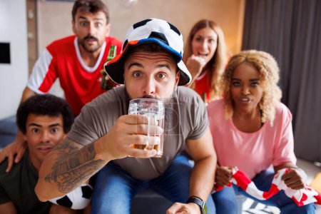 Foto de Grupo de amigos alegres fanáticos del fútbol que se divierten animando, bebiendo cerveza y viendo el juego del campeonato mundial en la televisión en casa - Imagen libre de derechos