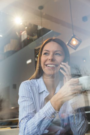 Retrato de una hermosa joven sentada en la mesa de un restaurante, mirando por la ventana, tomando café y hablando por teléfono