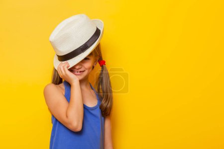 Téléchargez les photos : Portrait de belle petite fille vêtue d'une robe bleue et d'un chapeau d'été, cachant son visage derrière le chapeau et regardant, isolée sur fond jaune - en image libre de droit