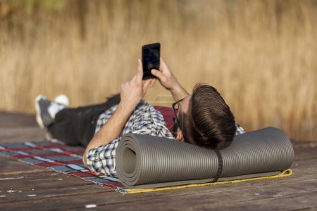 Téléchargez les photos : Homme allongé sur les quais du lac, relaxant après une longue journée de randonnée dans la nature, à l'aide d'un téléphone intelligent - en image libre de droit