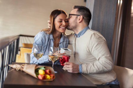 Photo for Couple on a date in a restaurant, celebrating anniversary, guy kissing a girl and giving her a present - Royalty Free Image