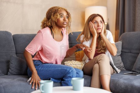 Téléchargez les photos : Deux amies s'amusent à passer du temps ensemble à la maison, à regarder des films d'horreur à la télé et à manger du pop-corn - en image libre de droit