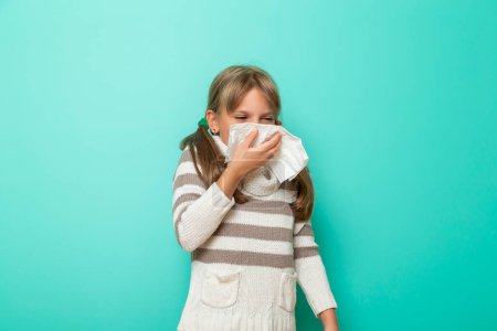 Téléchargez les photos : Portrait d'une petite fille ayant la grippe, se mouchant et s'essuyant avec des mouchoirs en papier isolés sur fond couleur menthe - en image libre de droit
