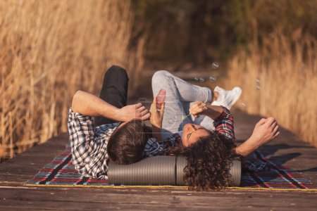 Foto de Pareja enamorada acostada en una manta de picnic en los muelles del lago, divirtiéndose mientras hace burbujas de jabón y disfruta de un hermoso día soleado de otoño en la naturaleza. - Imagen libre de derechos