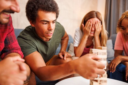Photo for Group of cheerful young friends having fun spending time together at home, playing jenga, take turns in removing blocks from a tower and stacking it on top of it - Royalty Free Image