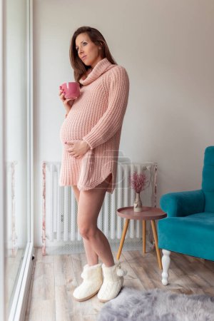 Foto de Retrato de una hermosa mujer embarazada parada junto a la ventana, bebiendo una taza de té caliente y relajándose en casa - Imagen libre de derechos