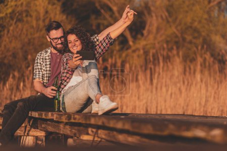 Téléchargez les photos : Couple assis sur les quais du lac, prendre un selfie avec un téléphone intelligent, boire de la bière et s'amuser - en image libre de droit