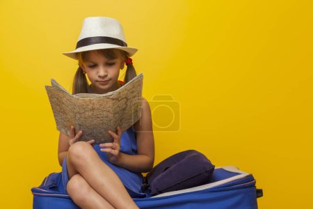 Foto de Hermosa niña con vestido azul de verano y sombrero sentado en su maleta y leyendo un mapa, preparándose para las vacaciones de verano temporada de viaje - Imagen libre de derechos