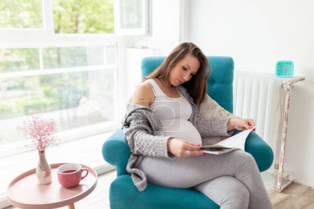 Photo for Beautiful pregnant woman sitting in an armchair by the window, relaxing at home, reading a magazine and drinking tea - Royalty Free Image
