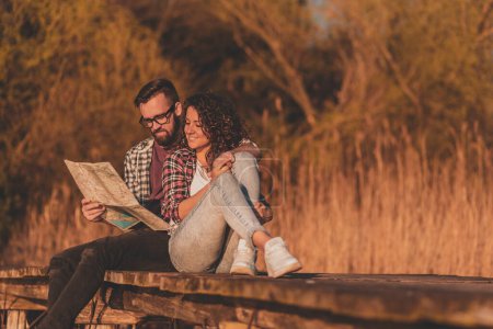 Foto de Hermosa pareja joven enamorada sentada en los muelles del lago, abrazando y leyendo un mapa, planeando una ruta de senderismo y disfrutando de un hermoso día de otoño en la naturaleza - Imagen libre de derechos