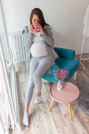 Foto de Retrato de una hermosa mujer embarazada sentada en un sillón cerca de la ventana, bebiendo una taza de té caliente y relajándose en casa - Imagen libre de derechos