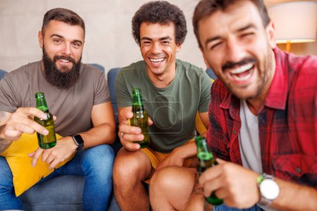 Foto de Grupo de amigos varones que se divierten pasando tiempo libre juntos en casa, viendo la televisión, comiendo palomitas de maíz y bebiendo cerveza - Imagen libre de derechos