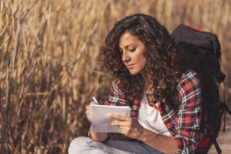 Téléchargez les photos : Belle jeune femme assise sur les quais du lac en bois, profitant d'une belle journée d'automne dans la nature et écrivant dans un carnet - en image libre de droit