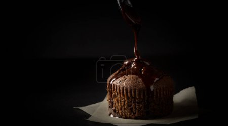 Téléchargez les photos : Muffin au chocolat recouvert de sauce au chocolat placé sur un morceau de papier sulfurisé sur fond noir - en image libre de droit